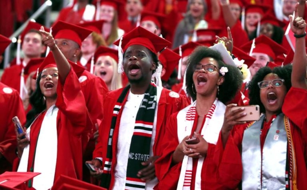 NC State students in red graduation attire in mid-celebration! They are cheering and yelling in a crowd.