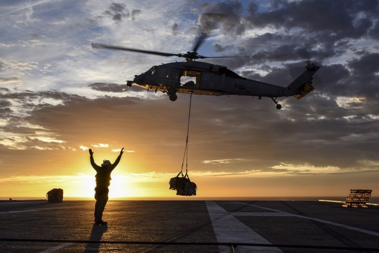 Military helicopter airlifting a supply drop with a crew member directing from the ground.