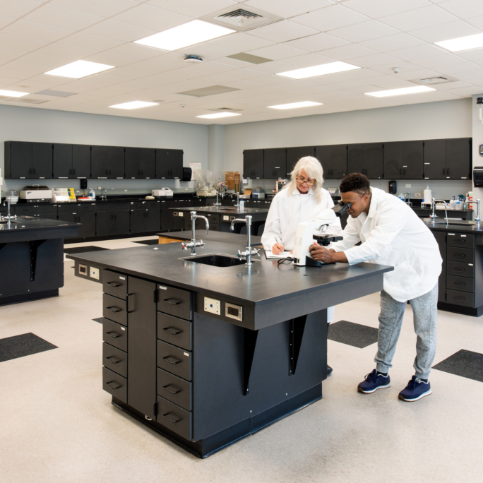 Two scientists working together. One scientist is carefully reviewing a sample under an electric microscope while the other observes and takes notes.