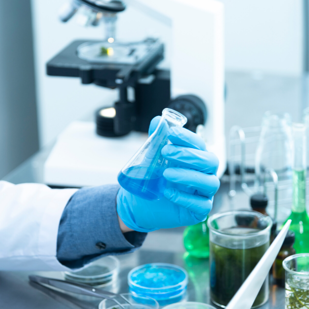 A pair of hands with medical gloves holding a glass beaker with a blue liquid solution in it. There is various lab equipment in the background, such as an electron microscope and other lab glassware.