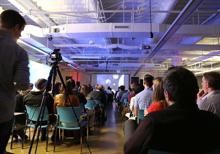 A large presentation with a seated crowd and a speaker distant at the front of the room. A videographer is quietly recording the presentation with a handheld video camera on a tripod.