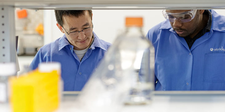 Two lab professionals working together. Lab equipment, such as glassware, is obscured in the foreground.